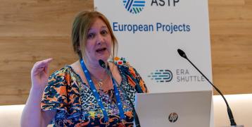 Laura MacDonald, CEO at ASTP Portrait of ASTP CEO Laura McDonald Speaking at ASTP Annual Conference. In the background there is a banner with all the EU projects ASTP is part of. Laura has a laptop with a michrophone in front of her, and she is looking directly in the camera.